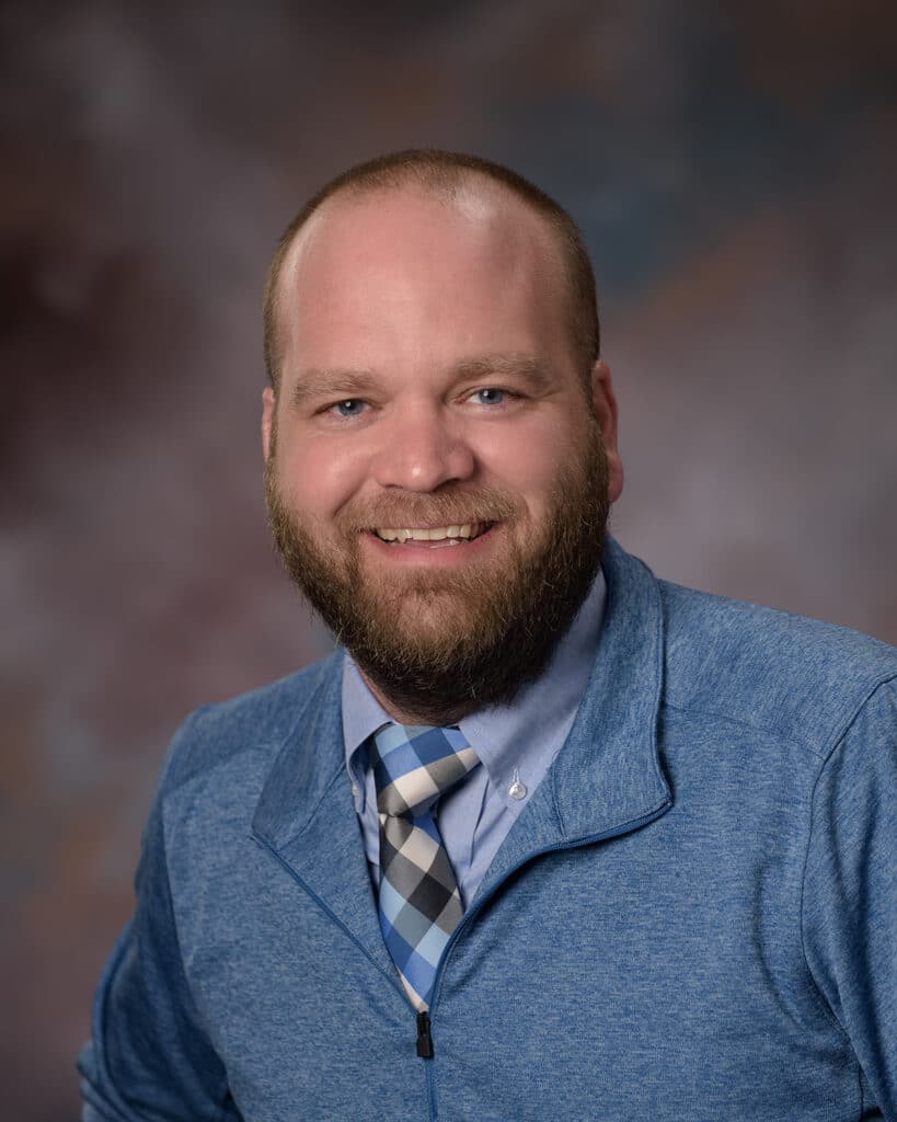 Ben Carlson, a bearded man with a shaved head, wearing a blue zip-up jacket over a dress shirt and plaid tie, smiles at the camera against a softly blurred brown and blue background.