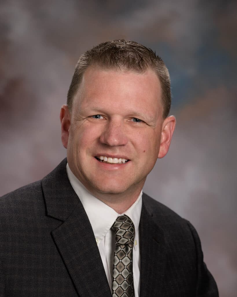 Travis Hansen, in a dark suit, white shirt, and patterned tie, smiles at the camera against a blurred, neutral background.