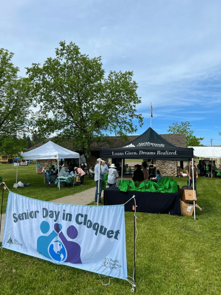 People gather outdoors at a park event under tents near a building. A banner in front reads "Senior Day in Cloquet," hosted by a local insurance agency. Tables with green bags are set up on the grass under a blue sky with trees in the background.