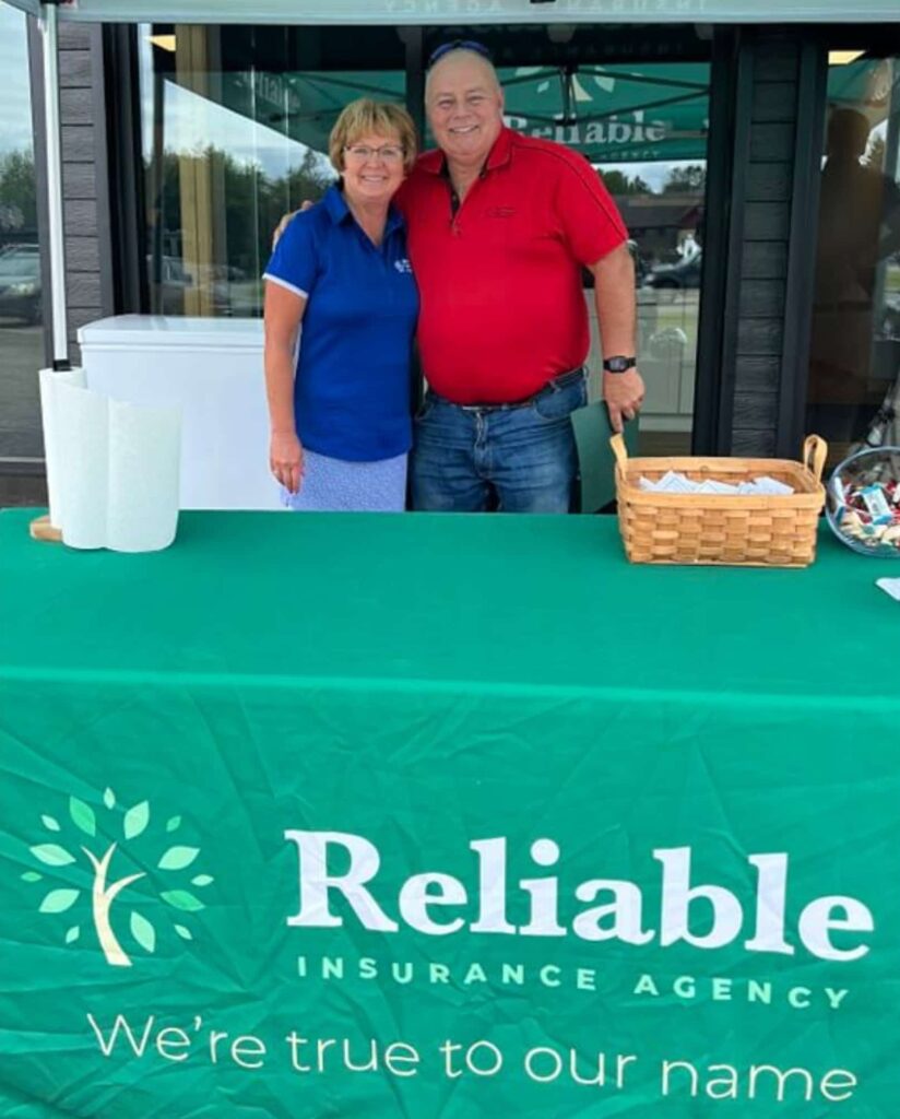 Two people stand behind a table covered with a green cloth that reads "Reliable Insurance Agency. We're true to our name." The pair is smiling, representing your local insurance agency, and a basket with items sits on the table.