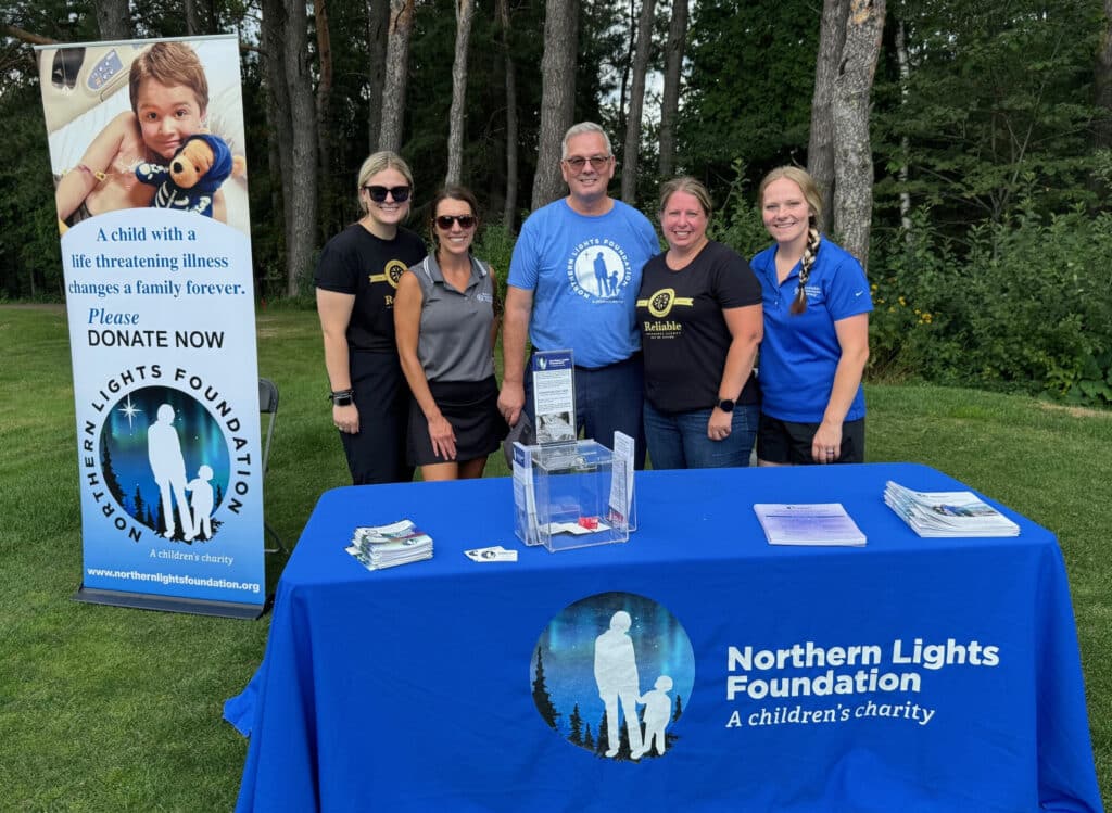 Five people stand smiling behind a blue table with a “Northern Lights Foundation” banner at an outdoor event, promoting a children’s charity. A Duluth Insurance Agency flyer about Personal Insurance and a donation box are displayed on the table.