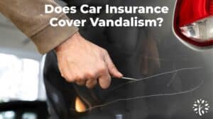 A person’s hand scratches the side of a dark-colored car with a sharp object; bold white text asks, “Does car insurance cover vandalism?” A small tree logo appears in the bottom right corner.