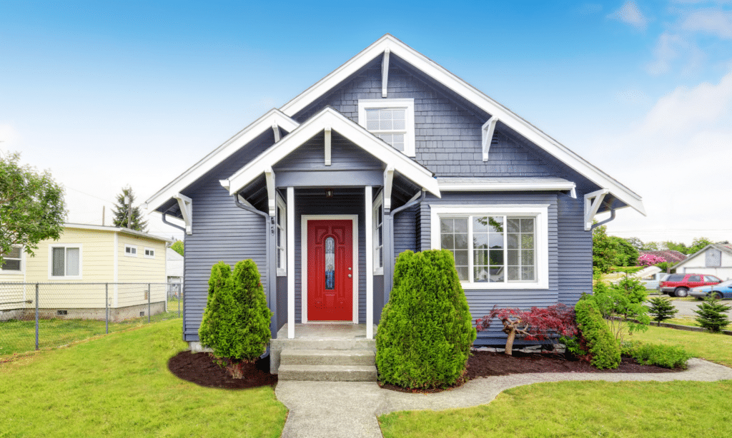 A charming blue house with white trim, a red front door, and covered porch. Well-kept shrubs and a green lawn frame the entrance on a sunny day with a clear blue sky.