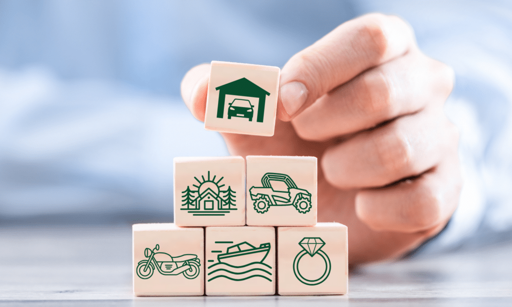 A hand stacks wooden blocks displaying icons of a house, car, motorcycle, boat, diamond ring, and off-road vehicle—Your Insurance Guide to understanding insurable assets.