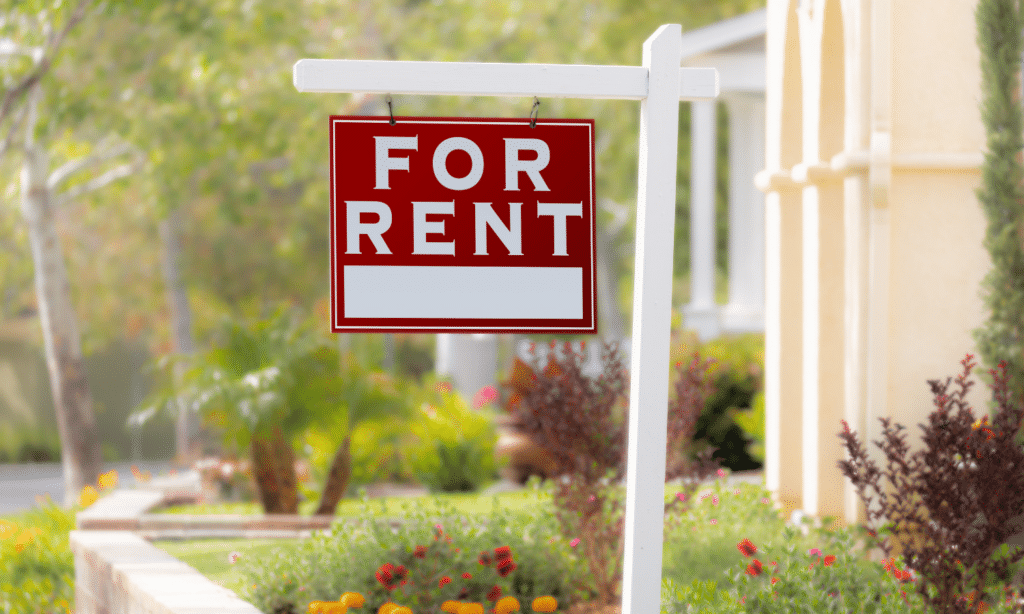 A red and white "For Rent" sign hangs from a white post in front of a house, with a garden of colorful flowers and greenery in the background—your insurance guide can help protect your new rental home.