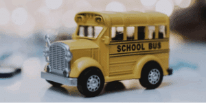 A close-up of a small yellow toy school bus with the words "SCHOOL BUS" on its side, placed on a smooth surface. The background is softly blurred with warm lights.
