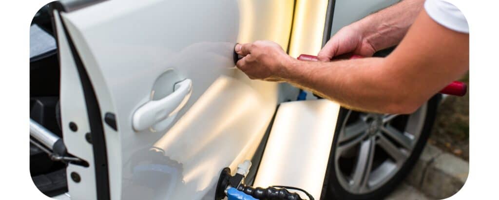 A person uses a tool to repair a dent in the door of a white car—possibly hail damage—while the car is parked on a street and equipment is visible nearby. Does Car Insurance Cover Hail Damage?.