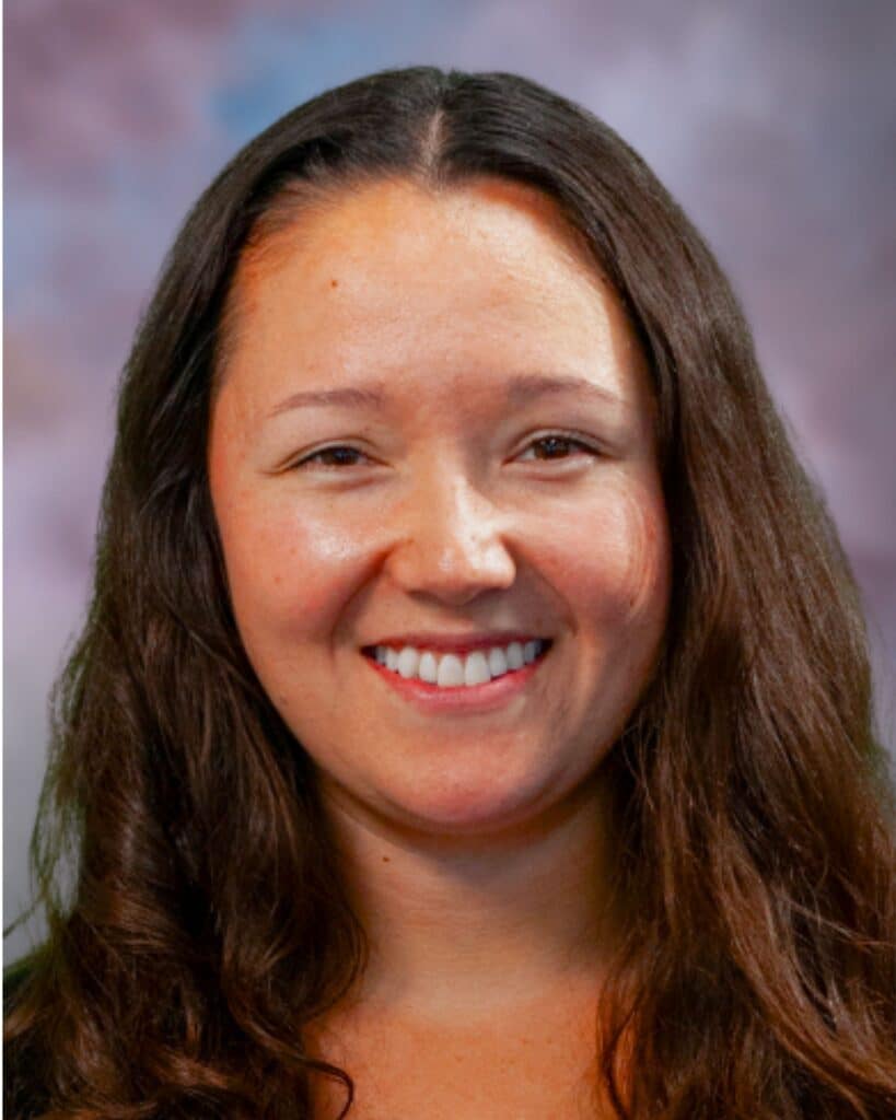 Adriana Wickett, a woman with long brown hair, smiles at the camera against a blurred background with shades of purple, blue, and gray.