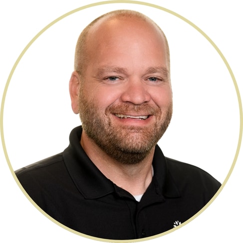Smiling man with a bald head and beard, wearing a black collared shirt, posed against a white background within a light circular border in the signature portrait style of Adriana Wickett.