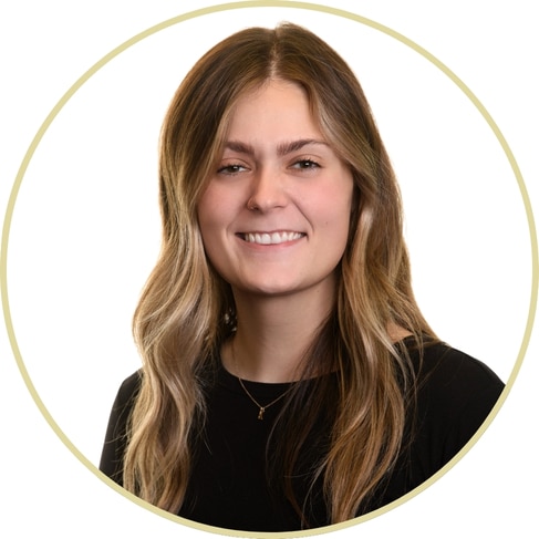 Adriana Wickett, a young woman with long, wavy light brown hair, wearing a black top and a delicate necklace, smiles in front of a white background with a circular border.