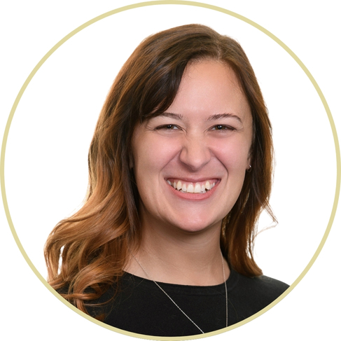 Adriana Wickett, a woman with long, wavy brown hair, smiles broadly. She is wearing a black top and a necklace, set against a plain white background with a light circular border around her.