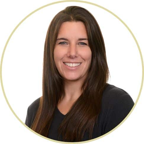 Adriana Wickett, a woman with long straight brown hair and blue eyes, is smiling at the camera. She is wearing a black top and is photographed against a plain white background within a circular frame.