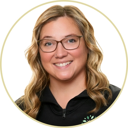 Smiling woman with wavy blond hair, glasses, and a nose piercing, wearing a black zip-up top. The image features Adriana Wickett framed by a light circular border against a white background.