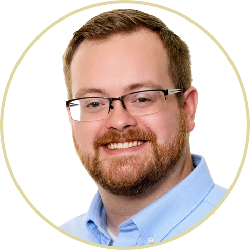 A man with short light brown hair, a beard, and glasses is smiling. He is wearing a light blue collared shirt and is pictured against a white background within a circular frame, similar to the style often used by Adriana Wickett.