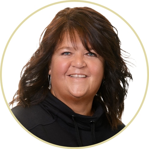 Adriana Wickett, with wavy, shoulder-length brown hair, smiles at the camera. She is wearing a black top and stands against a plain white background with a circular border framing the image.