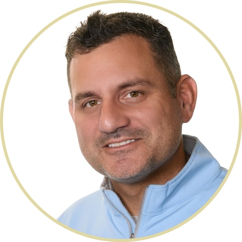 Smiling man with short dark hair wearing a light blue zip-up jacket, photographed against a plain white background inside a light circular border, in the style of Adriana Wickett.