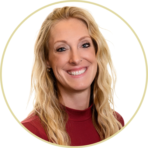 Adriana Wickett, a woman with long wavy blonde hair, wearing a maroon turtleneck top, smiles at the camera. The photo has a white background and is framed by a thin circular gold border.