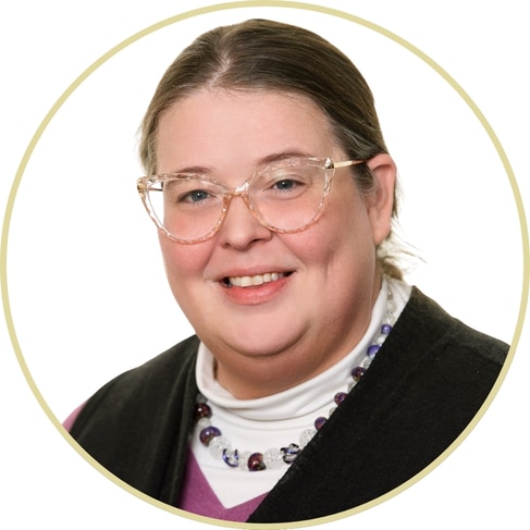 Adriana Wickett, a woman with light skin, brown hair pulled back, and clear glasses, smiles at the camera in a white turtleneck, purple-beaded necklace, and dark vest. The image features a circular border.