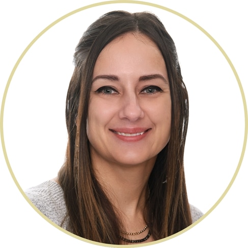 Adriana Wickett, a woman with long brown hair and blue eyes, smiles at the camera. She is wearing a light-colored sweater and a necklace, set against a plain white background with a circular border.