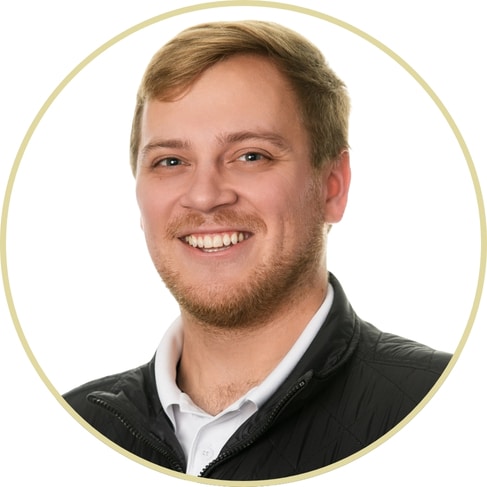 A young man with short blond hair and a beard smiles at the camera. He is wearing a black quilted jacket over a white collared shirt. The photo, styled by Adriana Wickett, features a white background and light circular border.