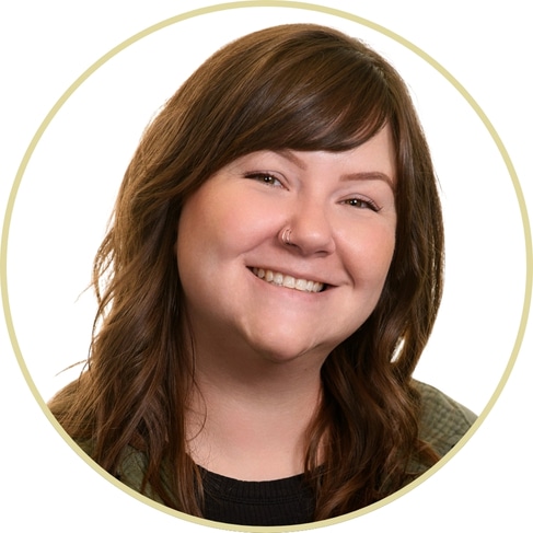 Adriana Wickett, a woman with long brown hair, wearing a green top and a nose ring, smiles at the camera against a plain white background, framed by a circular border.