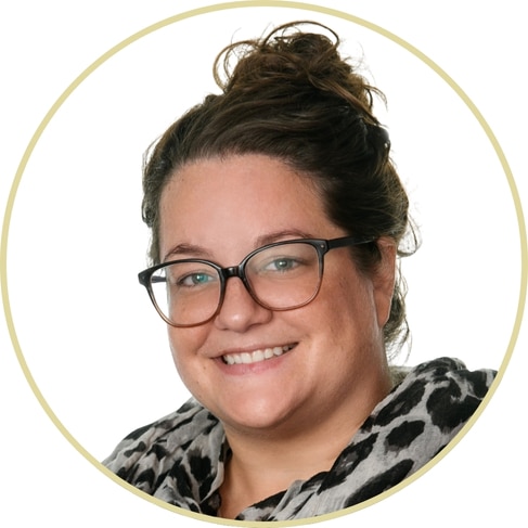 A smiling woman, Adriana Wickett, with brown hair in a messy bun, wears large black glasses and a black-and-white patterned top, pictured against a white background with a light circular border.