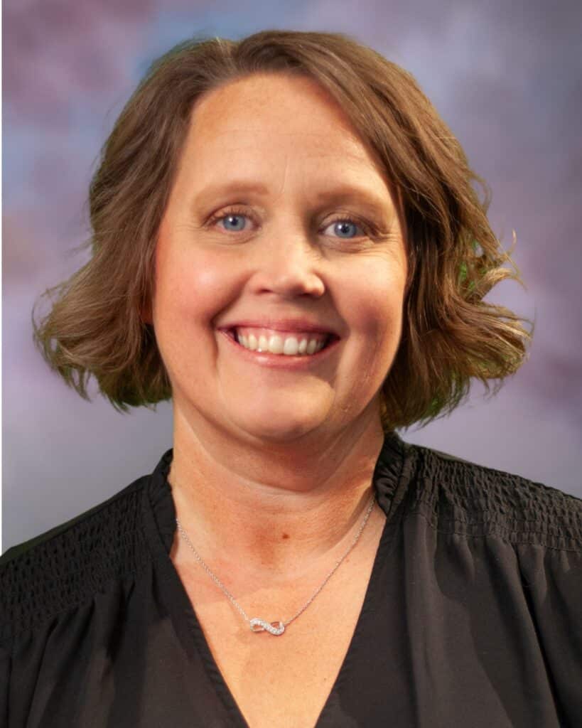 Sonja Carlson, a woman with short, wavy brown hair, smiles at the camera. She wears a black top and a silver infinity necklace, set against a softly blurred background in pastel colors.