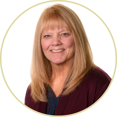 Adriana Wickett, with straight, shoulder-length blonde hair, smiles at the camera. She is wearing a maroon cardigan over a patterned blouse, set against a white background with a circular border.