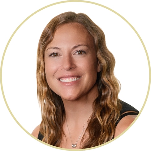 Adriana Wickett, with long, wavy light brown hair and a black sleeveless top, smiles at the camera. She wears a silver necklace and is shown against a white background within a circular border.