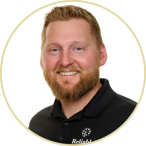 Smiling man with short, light brown hair and a beard, wearing a black collared shirt with a white logo, shown in a circular frame on a white background, reminiscent of a professional portrait by Adriana Wickett.