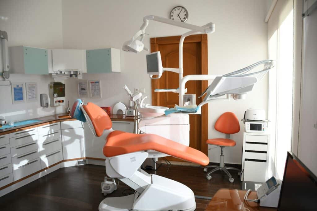 A modern dental clinic room with an orange dental chair, overhead light, dental equipment, a clock on the wall, cabinets, and a large window letting in natural light.