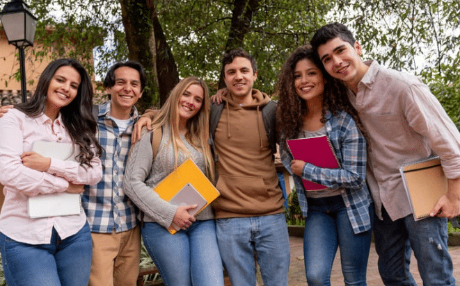 Six smiling young adults stand close together outdoors, holding notebooks and backpacks, with trees and a lamp post in the background, suggesting a cheerful campus or park setting.