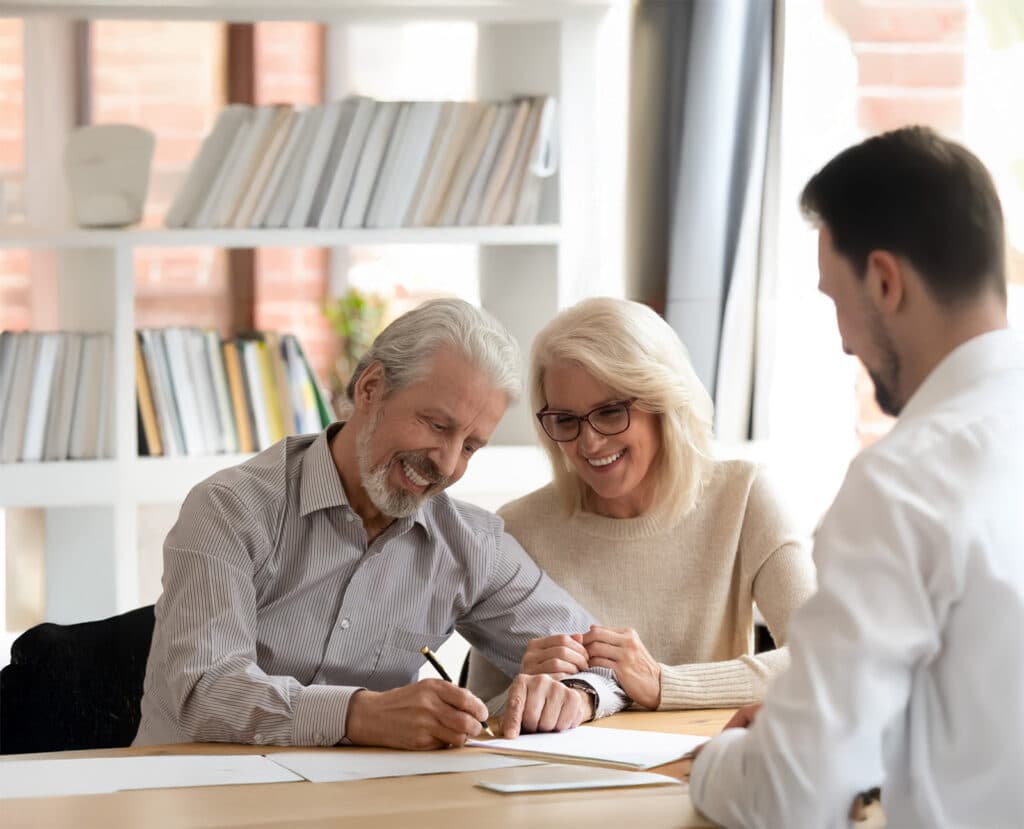 An older man smiles as he signs a Medicare document, with an older woman beside him and a younger man in a white shirt across the table in a bright office lined with bookshelves.