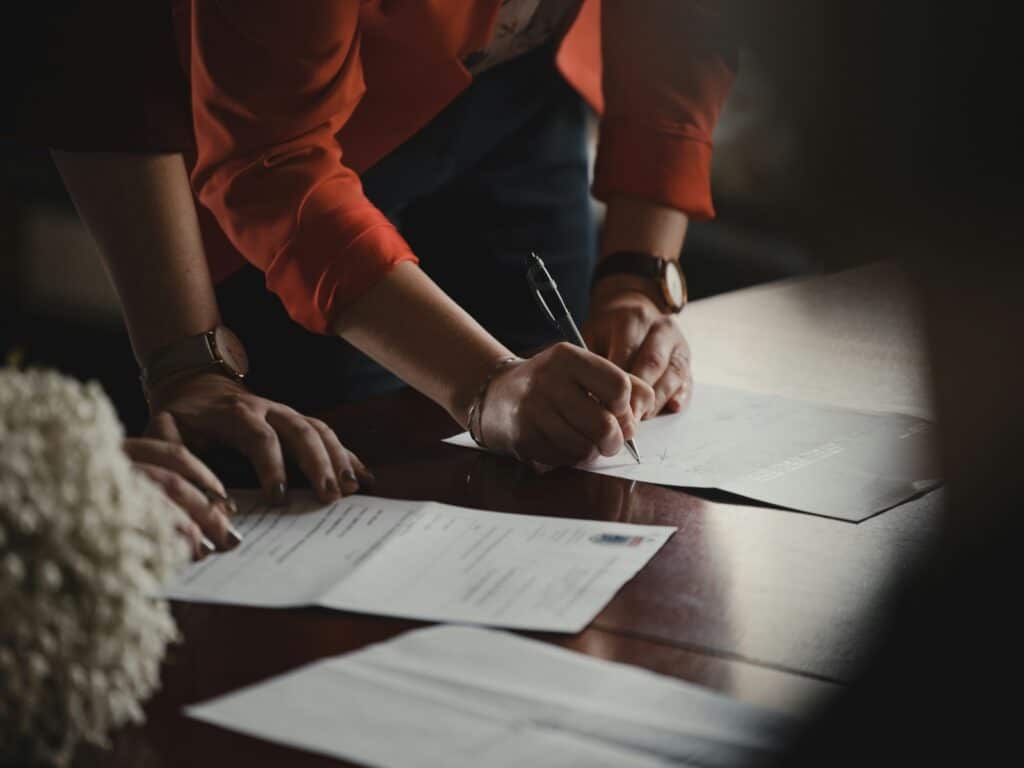 A person in a red jacket is leaning over a table, signing a Medicare document with a pen. Other papers and hands are visible on the table, suggesting a formal or official signing process.