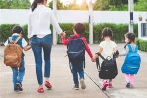 An adult and four young children with backpacks walk hand in hand on a paved path outdoors, embodying Back-to-school bus safety: A Family Guide for Northland Kids & Parents, surrounded by greenery as they head away from the camera.