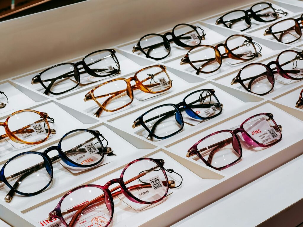 Rows of eyeglasses in various colors and styles are displayed on a white tray, each pair labeled with tags and QR codes.