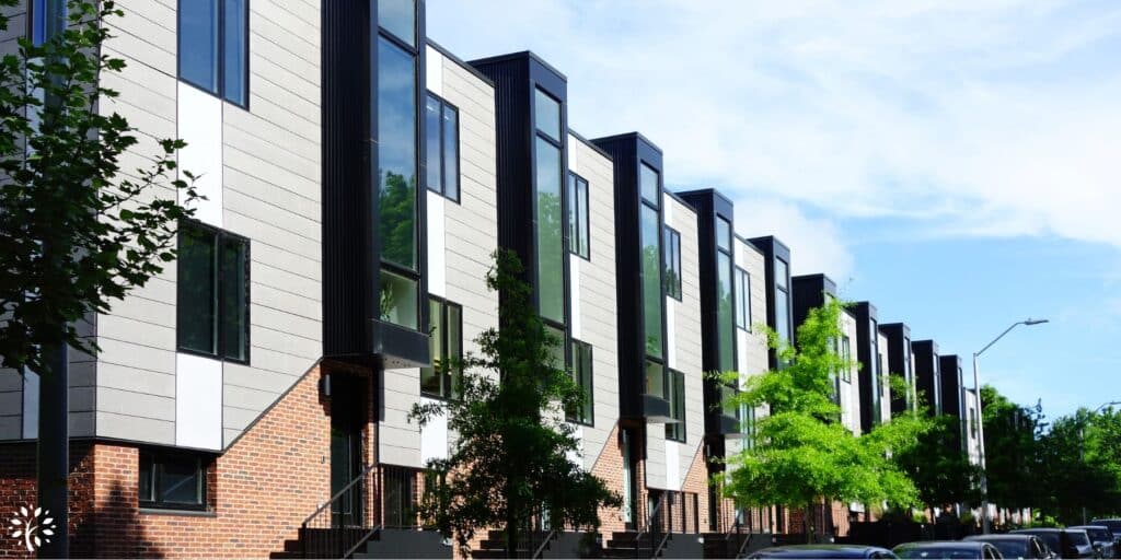 Modern row of three-story townhouses with large vertical windows, brick ground level, gray and white siding above, and trees lining the sidewalk on a sunny day. Cars are parked along the street.