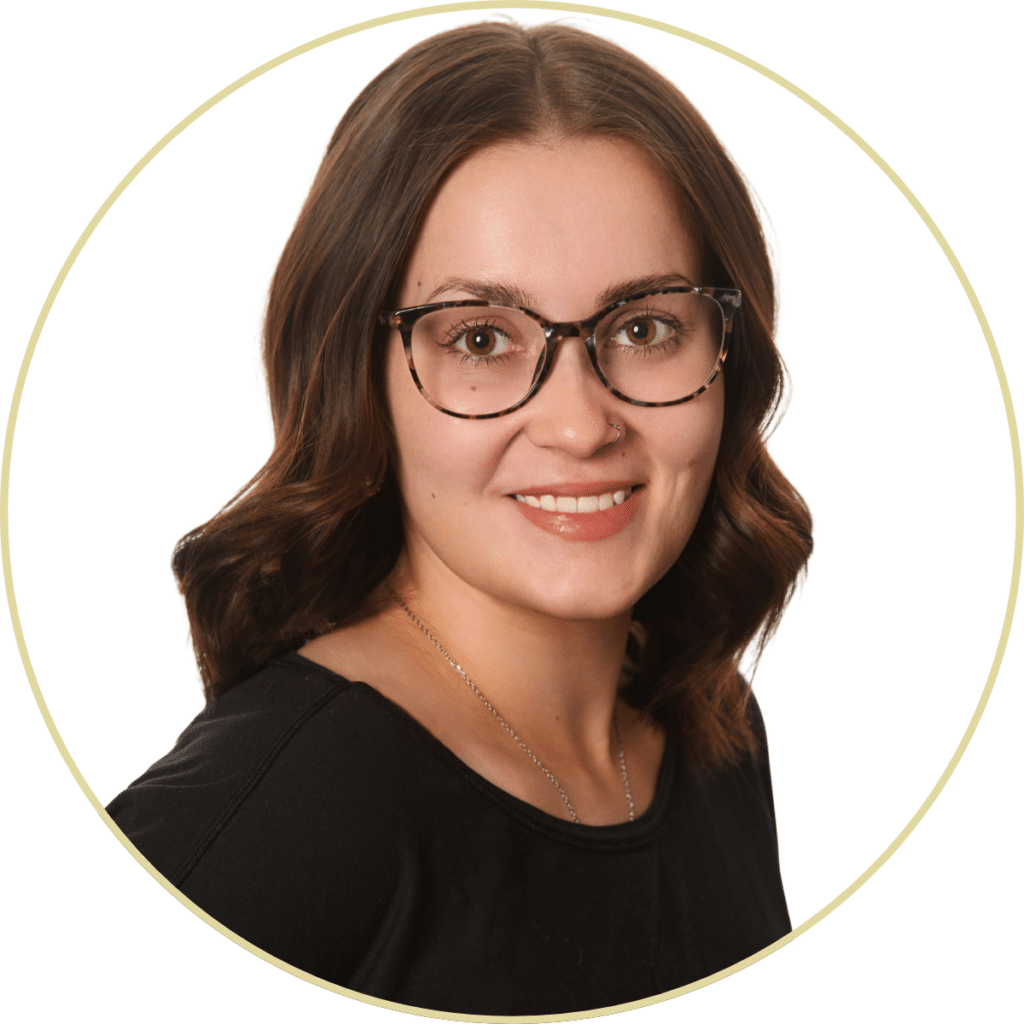 A woman with wavy brown hair, wearing tortoiseshell glasses and a black top, smiles at the camera against a white background—Lacy Lekander exudes warmth and confidence.