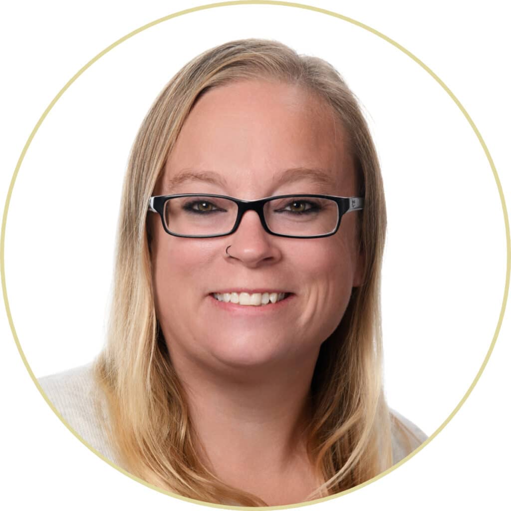 Smiling person with long blonde hair and glasses, wearing a light top, in front of a white background.